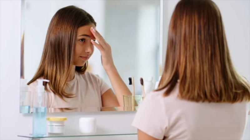 A Young Girl Is Inspecting Her Forehead in The Mirror, Likely Looking at An Acne Pustule