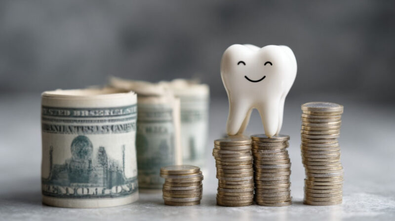 Stacks of coins and rolled-up dollar bills with a smiling tooth figurine on top, symbolizing dental expenses