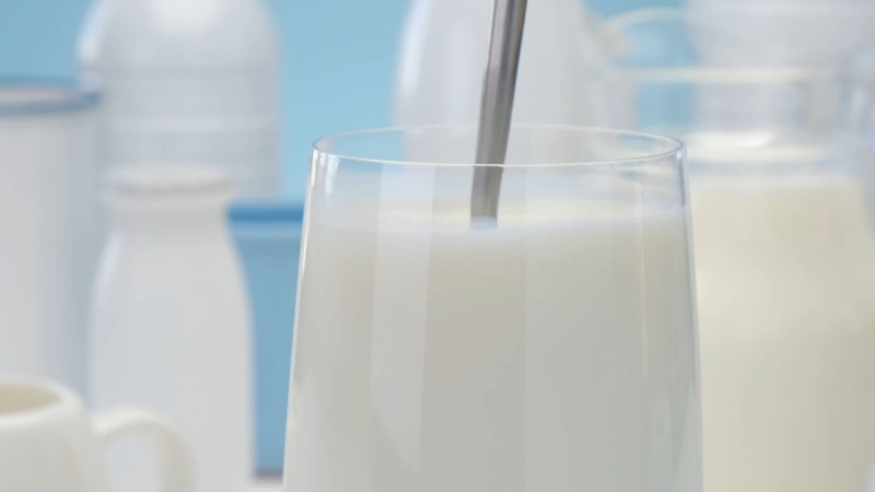 A close-up of a glass of dairy milk with a metal spoon and bottles in the background