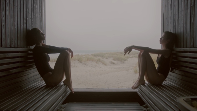 Two women sit across from each other in a heat room with an open view of sand and fog outside