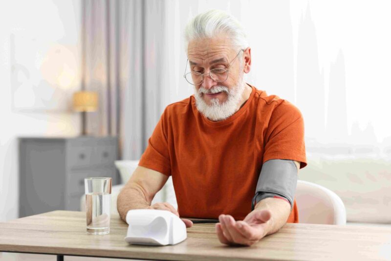 Older man sitting at a table using a blood pressure monitor with cuff on his arm