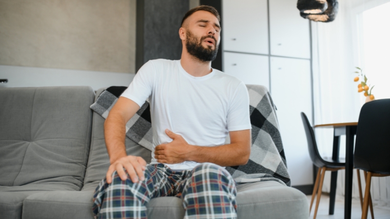 Man sitting on a couch holding his stomach due to gas and bloating