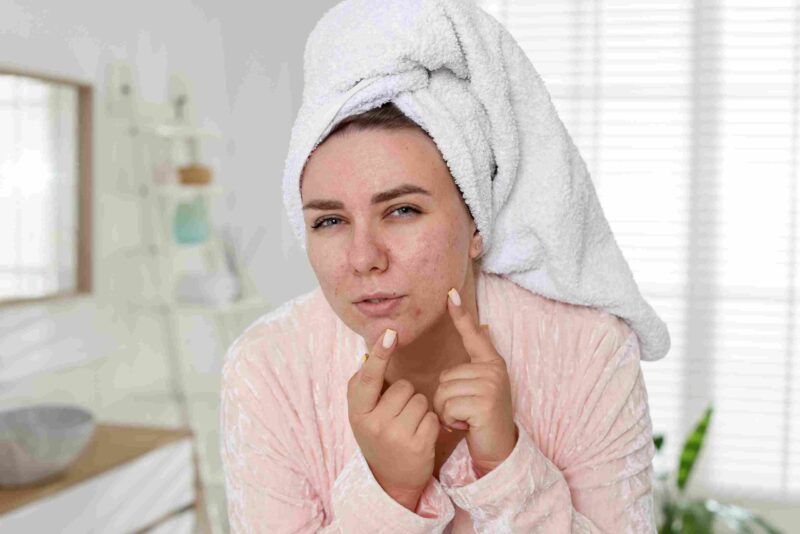 Woman with a towel on her head examining acne on her face in a bathroom mirror