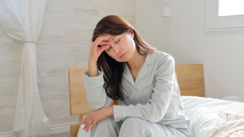 Woman sitting on bed holding her head, showing signs of waking up tired every morning